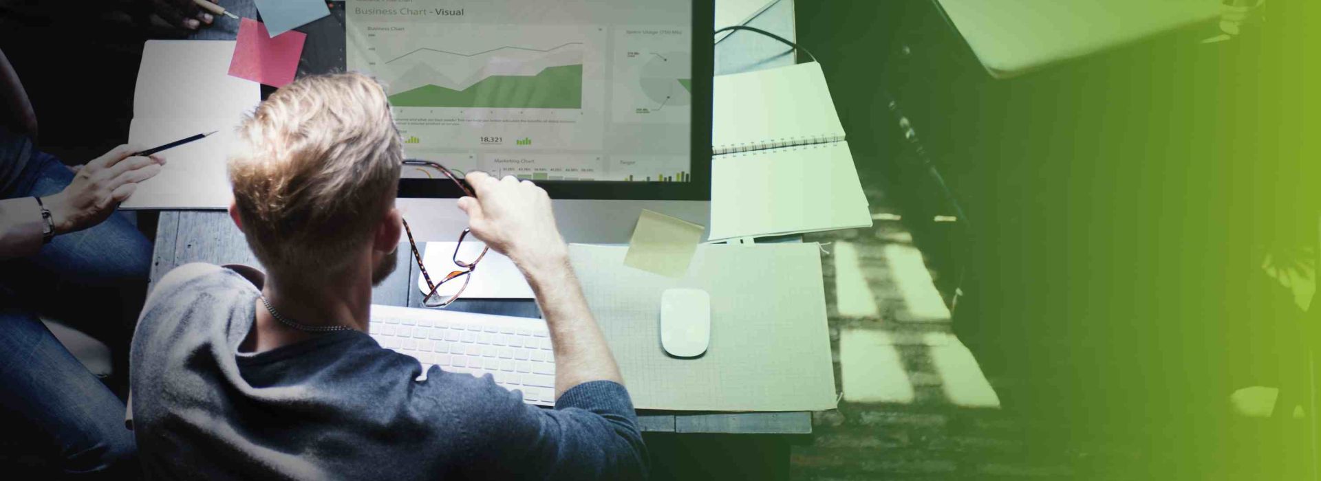 Ein blonder Mann hat seine Brille in der Hand und sitzt an einem Schreibtisch vor einem Computer