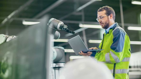 A man in a yellow and green safety jacket works on a robot with a laptop in his hand