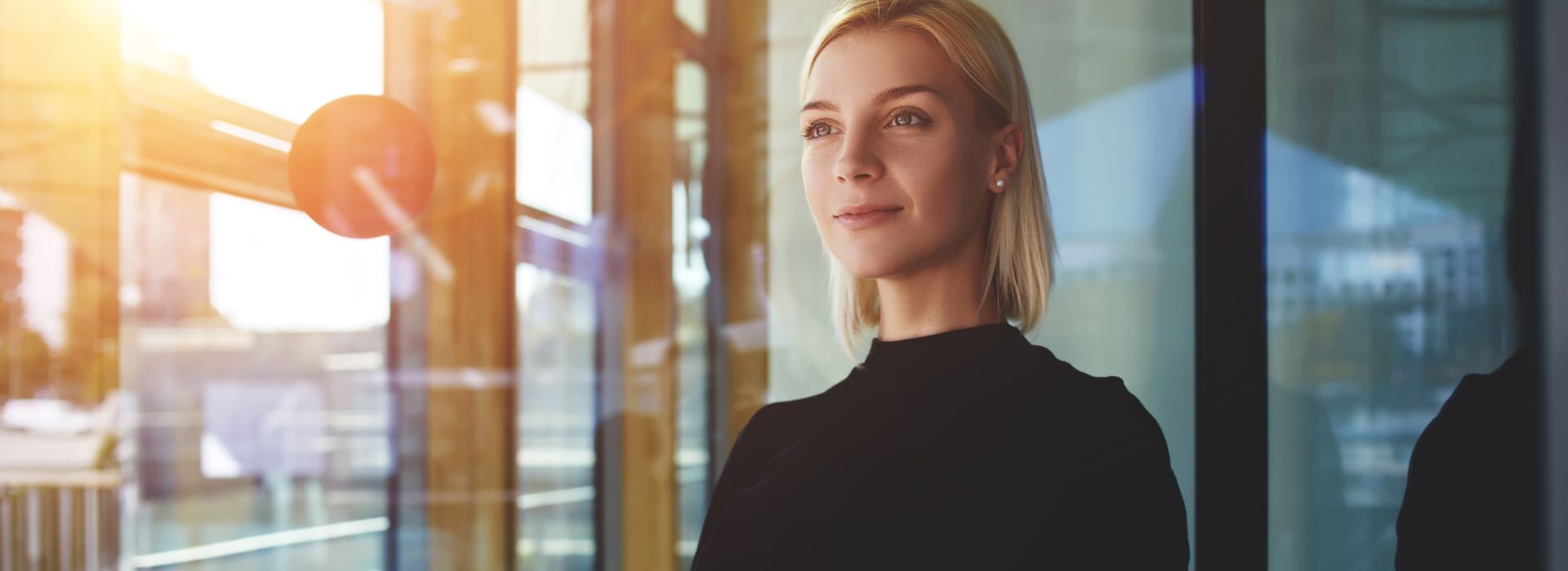 A woman with blonde hair and a black sweater stands in front of windows with a tablet in her hand.