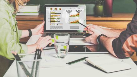 A woman and a man sit opposite each other at a desk and look at a laptop.