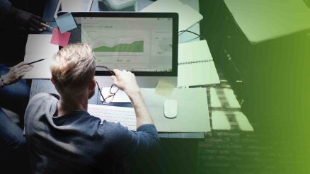 A blond man has his glasses in his hand and is sitting at a desk in front of a computer.